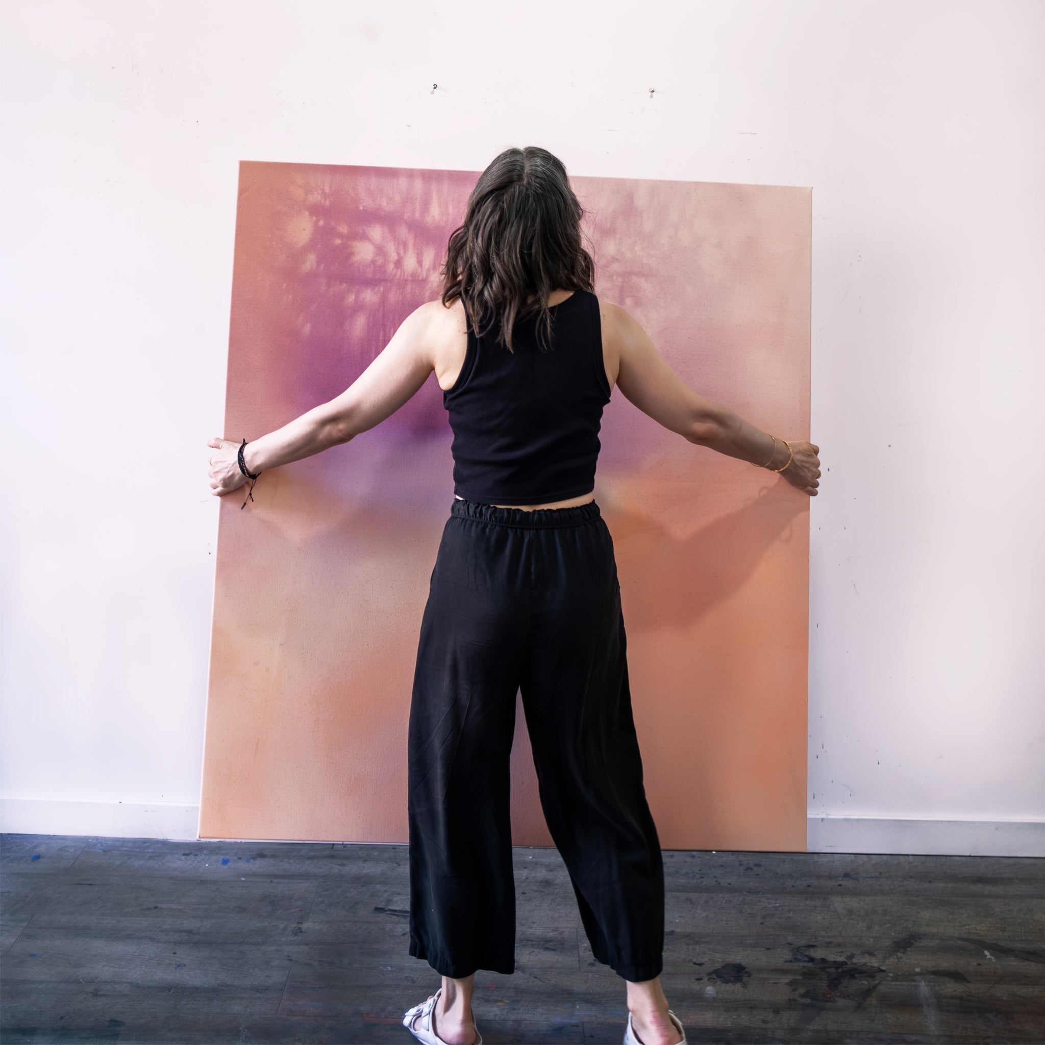 The artist stands with her back towards us. She is holding a large canvas from her Slow Burn series and is about to hang it on the wall.