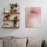 An abstract painting in washes of purple, yellow, pink, and blue hangs next to a shelving system of plants and above a couch with lots of pillows.